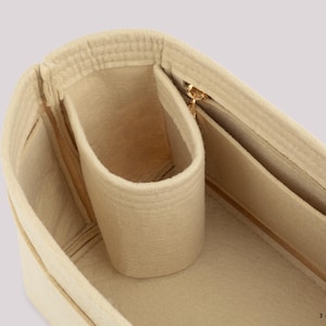 May include: Beige felt handbag organizer with multiple compartments and a gold-toned zipper. The organizer features a cylindrical pocket and several rectangular pockets for storing items. The interior is a light beige color.