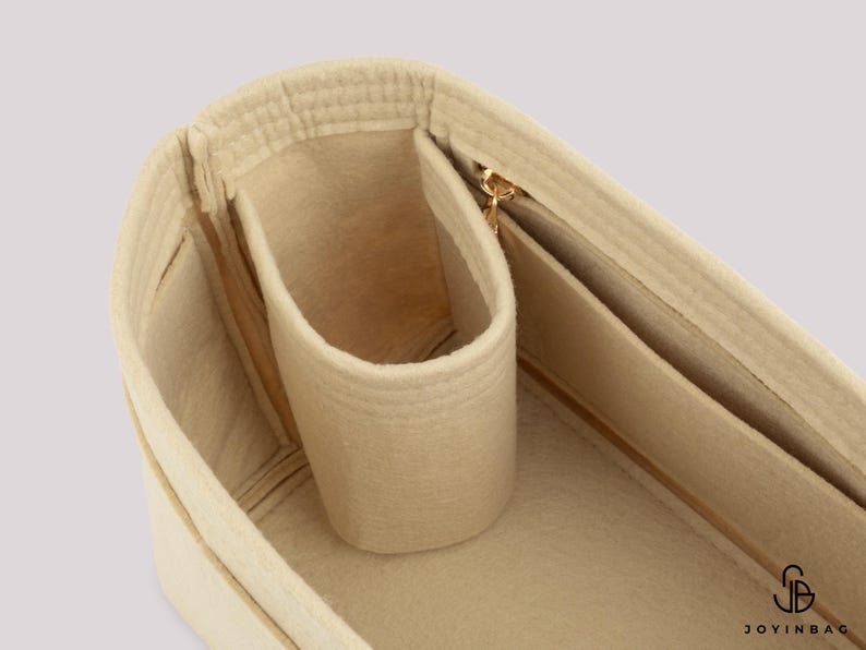 May include: Beige felt handbag organizer with multiple compartments and a gold-toned zipper. The organizer features a cylindrical pocket and several rectangular pockets for storing items. The interior is a light beige color.