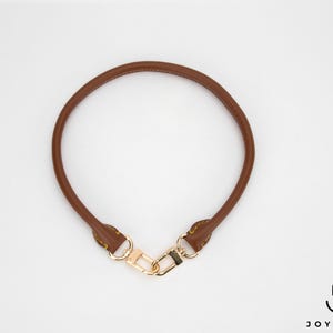 May include: A brown leather handbag handle with gold-coloured hardware. The handle is circular and has a clasp for easy attachment. The handle is a replacement part for a purse or handbag.