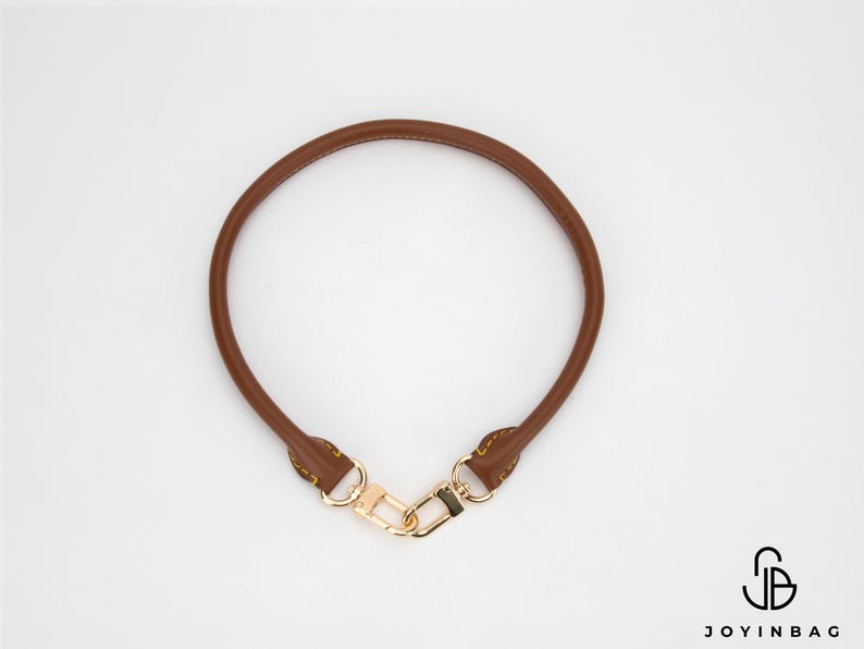 May include: A brown leather handbag handle with gold-coloured hardware. The handle is circular and has a clasp for easy attachment. The handle is a replacement part for a purse or handbag.