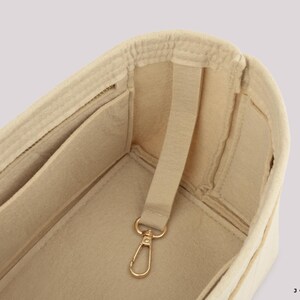 May include: Beige felt bag organizer with multiple pockets and a gold-colored clasp. The organizer has a structured design with a strap for easy handling. The interior features several compartments for storage.