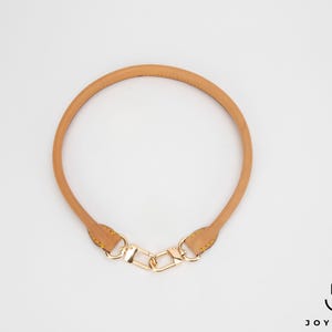 May include: A tan leather bag handle with gold-coloured hardware. The handle is circular and features a clasp at each end for easy attachment to a bag. The hardware includes a chain-link design.