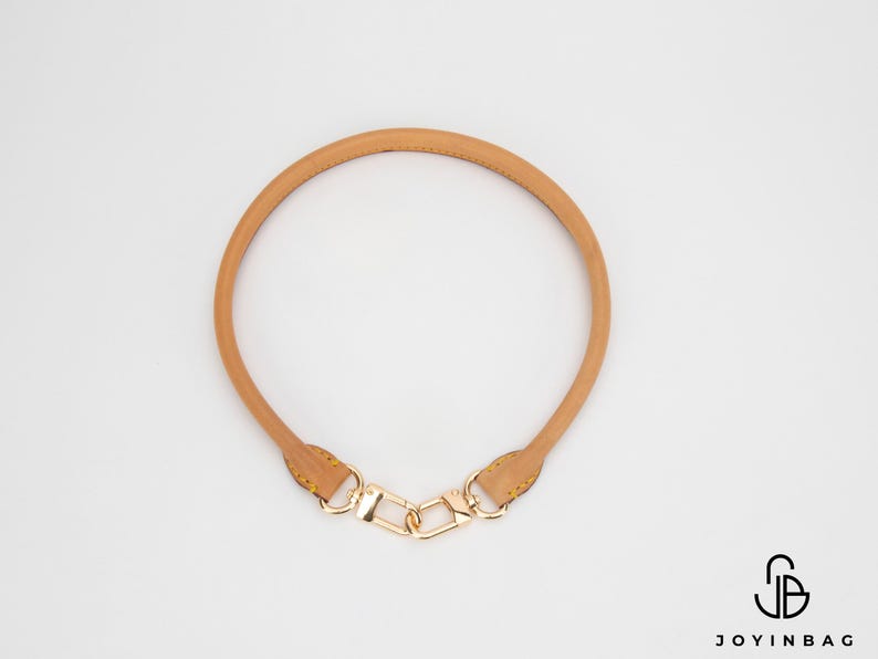 May include: A tan leather bag handle with gold-coloured hardware. The handle is circular and features a clasp at each end for easy attachment to a bag. The hardware includes a chain-link design.