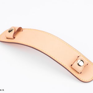 Puede incluir: Un asa de cuero marr&oacute;n claro con detalles en metal dorado. El asa tiene un borde cosido y est&aacute; dise&ntilde;ada para ser fijada a un bolso.
