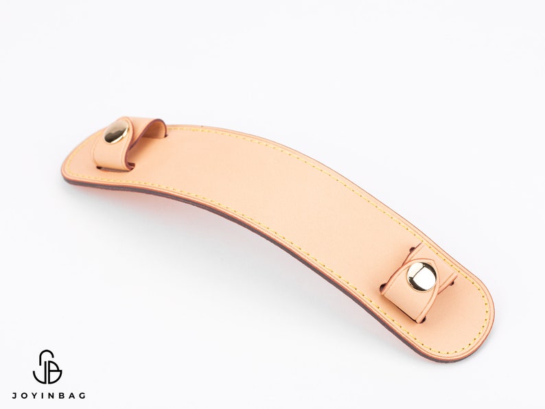 Puede incluir: Un asa de cuero marr&oacute;n claro con detalles en metal dorado. El asa tiene un borde cosido y est&aacute; dise&ntilde;ada para ser fijada a un bolso.