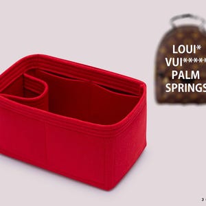 Op de afbeelding: Een rechthoekige tasorganizer van rood vilt met meerdere binnenvakken en een cilindervormig zijvak. Op de achtergrond is een wazige bruine rugzak met patroon zichtbaar met de tekst LOUI* VUI***** PALM SPRINGS.