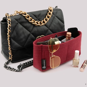May include: A black quilted handbag with a gold chain strap positioned behind a rectangular burgundy felt purse organiser. The organiser contains gold-rimmed sunglasses, a small bottle, and a wallet, with nail polish and lipstick nearby.