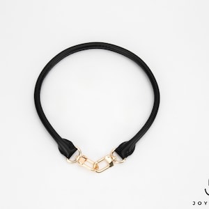 May include: A black leather bag handle with gold-coloured metal clasps. The handle is circular and appears to be made of a soft, flexible material. The clasps are rectangular and connect the handle ends.