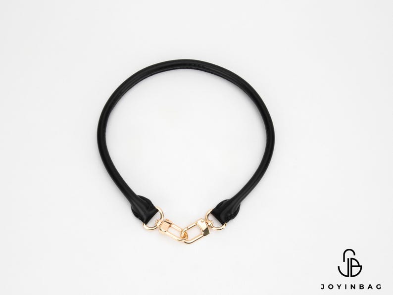 May include: A black leather bag handle with gold-coloured metal clasps. The handle is circular and appears to be made of a soft, flexible material. The clasps are rectangular and connect the handle ends.