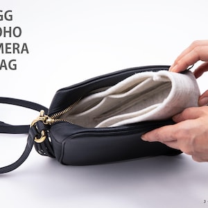 May include: A black leather camera bag with gold-tone hardware and a long shoulder strap. Inside the bag is a removable off-white felt organizer insert with multiple compartments. The text GG SOHO CAMERA BAG is printed in the upper left corner.