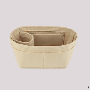 May include: Beige bag organizer with multiple compartments and a zippered pocket. The organizer is designed to fit inside a handbag, providing structure and organization. The product is made of a soft, felt-like material.
