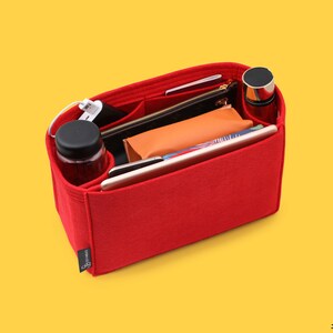 May include: Red felt handbag organizer with multiple compartments and pockets. The organizer is filled with various items, including a water bottle, phone, wallet, and a small cosmetic bag.