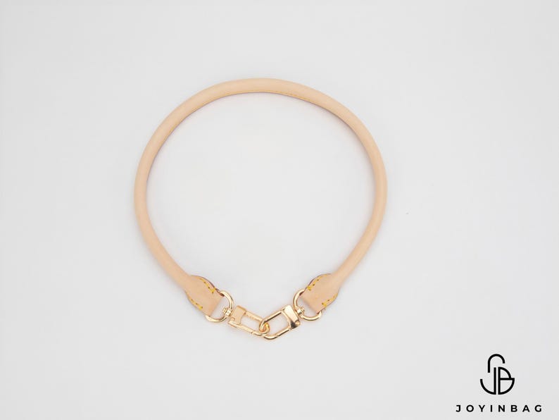 May include: A light tan leather bag handle with gold-coloured hardware. The handle is circular and has a clasp for easy attachment. The image is on a white background.