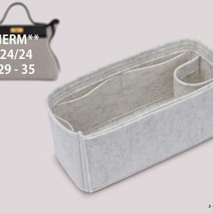 May include: A light grey felt handbag organiser insert with multiple interior compartments and a structured rectangular shape. The insert features a small black side tag with the text JOYINBAG and a graphic of a handbag with text HERM** 24/24 29-35.