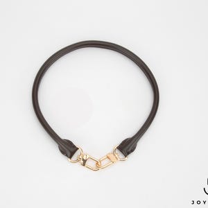 May include: A dark brown leather bag handle with gold-coloured clasps. The handle is circular and appears to be made of a smooth, durable material. The clasps are connected by a gold-coloured clasp.