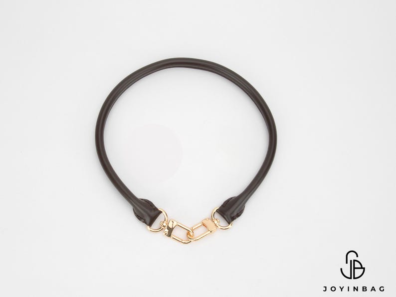 May include: A dark brown leather bag handle with gold-coloured clasps. The handle is circular and appears to be made of a smooth, durable material. The clasps are connected by a gold-coloured clasp.