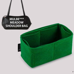 以下が含まれることがあります： 複数のコンパートメントを備えた明るい緑色のフェルトバッグオーガナイザー。 オーガナイザーはハンドバッグの内側に収まるように設計されています。 背景には「MULBE*** MEADOW SHOULDER BAG」の文字が入った黒いショルダーバッグがあります。
