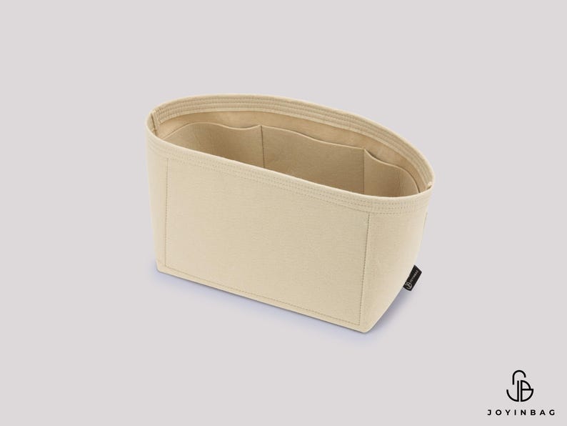 May include: Beige handbag organizer with multiple interior pockets. The rectangular organizer is made of a soft, light-colored fabric. The interior features several pockets for organization. A small black tag is attached to the side.