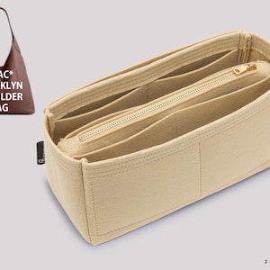 Può includere: Un organizer per borsa in feltro beige con molteplici scomparti interni e una tasca centrale con cerniera. L'inserto è progettato per una borsa a spalla marrone etichettata COAC* BROOKLYN SHOULDER BAG. Una piccola etichetta nera è fissata sul lato.