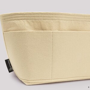 May include: Beige felt bag organizer with multiple compartments and stitched details. The organizer has a rectangular shape with a curved bottom and a small black tag with the text "JOYINBAG".