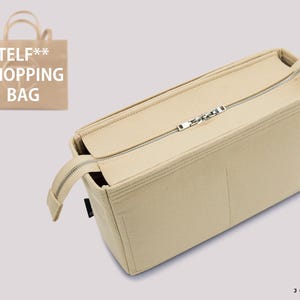 May include: A beige felt bag organizer insert featuring a rectangular shape, multiple interior compartments, and a silver-toned metal zipper closure across the top. A small graphic in the corner shows a beige shopping bag with the text TELF** SHOPPING BAG.