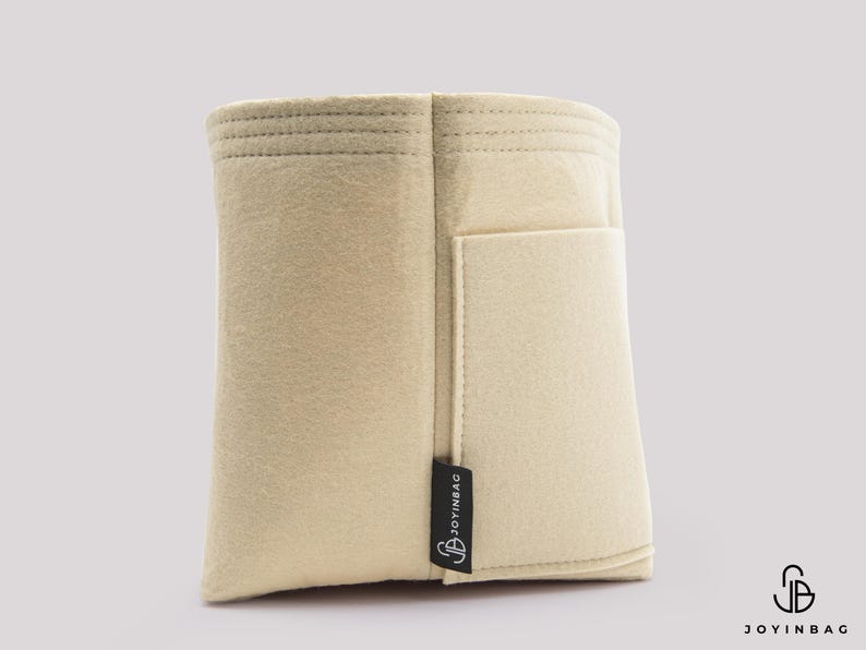 May include: A beige felt bag organizer with a rectangular shape and a small pocket on the side. The organizer has a stitched top and a black tag that reads "JOYINBAG". The bag is designed to help organize items inside a larger bag.