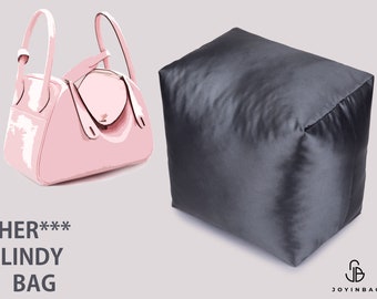 May include: A pink leather handbag with a top flap closure and two handles. The bag is sitting on a white surface. The text "HER*** LINDY BAG" is below the bag.