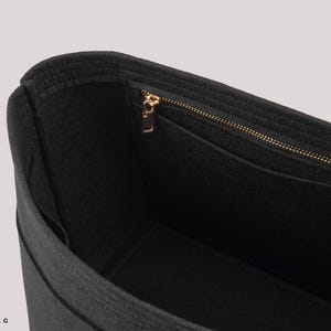 May include: Black felt handbag organizer with a gold-colored zipper and multiple pockets. The interior is visible, showcasing the organizational structure. The bag is designed to fit inside a larger tote or handbag.