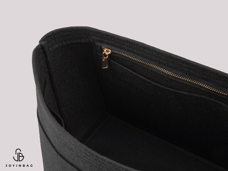 May include: Black felt handbag organizer with a gold-colored zipper and multiple pockets. The interior is visible, showcasing the organizational structure. The bag is designed to fit inside a larger tote or handbag.