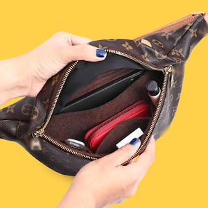 Könnte beinhalten: Ein brauner Taschenorganizer aus Filz in einer braunen Gürteltasche mit Monogramm-Muster. Der Einsatz verfügt über mehrere Fächer, die ein schwarzes Smartphone, eine rote Reißverschlussbrieftasche, ein weißes Objekt und eine schwarze Tube enthalten.