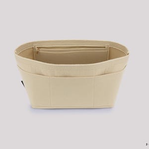 May include: Beige felt handbag organizer with multiple pockets and a zippered compartment. The organizer is designed to fit inside a handbag, providing structure and organization. The product is made of a soft, neutral-colored felt material.