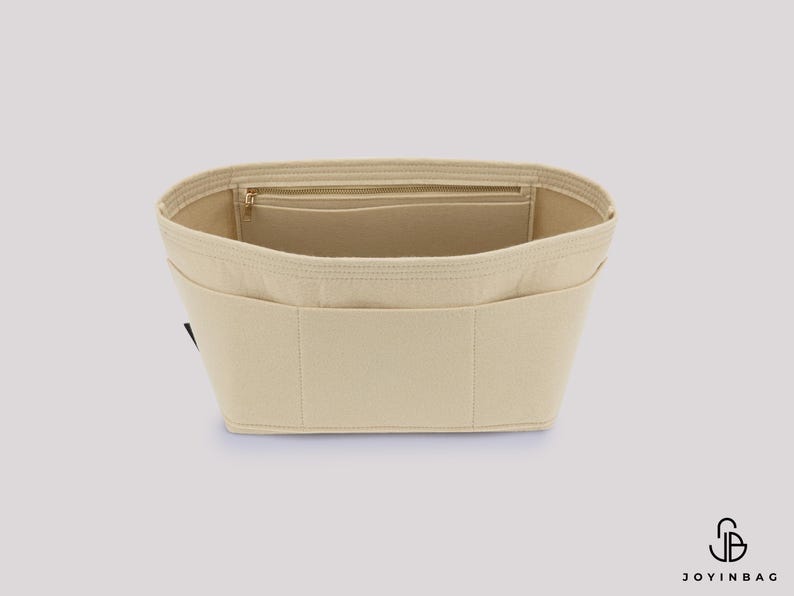 May include: Beige felt handbag organizer with multiple pockets and a zippered compartment. The organizer is designed to fit inside a handbag, providing structure and organization. The product is made of a soft, neutral-colored felt material.