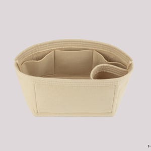 May include: Beige handbag organizer with multiple compartments and pockets. The organizer is designed to fit inside a larger bag, providing structure and organization. It has a rectangular shape with a curved top and is made of a soft, felt-like material.