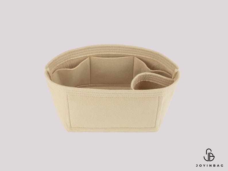May include: Beige handbag organizer with multiple compartments and pockets. The organizer is designed to fit inside a larger bag, providing structure and organization. It has a rectangular shape with a curved top and is made of a soft, felt-like material.