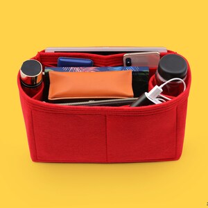 May include: Red felt bag organizer with multiple compartments. The organizer is filled with various items, including a phone, a water bottle, a power bank, and a notebook.