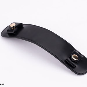 Puede incluir: Asa de cuero negro con herrajes de metal dorado. El asa es curva y tiene dos botones de metal dorado en cada extremo.