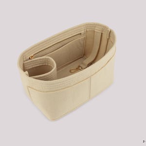 May include: Beige bag organizer with multiple compartments, including a zippered pocket and a small cylindrical pocket. Features a gold-colored zipper and clasp. The organizer is made of a soft, felt-like material.