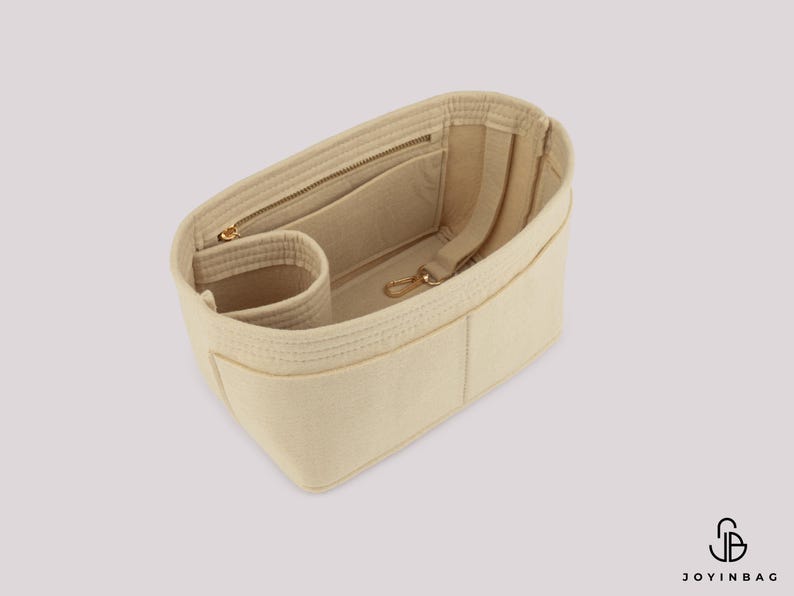 May include: Beige bag organizer with multiple compartments, including a zippered pocket and a small cylindrical pocket. Features a gold-colored zipper and clasp. The organizer is made of a soft, felt-like material.