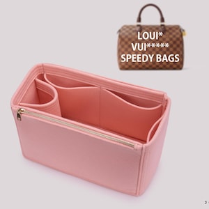 Peut inclure: Un organisateur de sac à main en feutre rose clair avec plusieurs compartiments intérieurs et une poche zippée dorée. En arrière-plan, un sac à main à carreaux marron est visible avec le texte LOUI* VUI***** SPEEDY BAGS imprimé au-dessus.