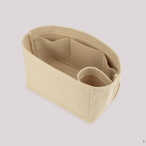 May include: Beige handbag organizer with multiple compartments and pockets. The interior features a large open space, several smaller pockets, and a cylindrical holder. The organizer is designed to keep items organized inside a handbag.