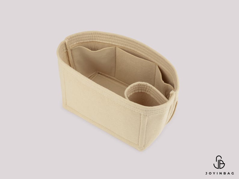 May include: Beige handbag organizer with multiple compartments and pockets. The interior features a large open space, several smaller pockets, and a cylindrical holder. The organizer is designed to keep items organized inside a handbag.