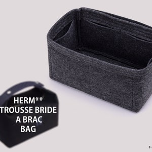 Op de afbeelding: Grijze feltas-organizer met meerdere vakken. De tas-organizer heeft een handvat en de tekst "HERM** TROUSSE BRIDE A BRAC BAG" gedrukt op de voorkant.