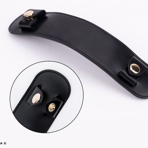 Puede incluir: Asa de cuero negro con detalles en metal dorado. El asa es curva y tiene dos peque&ntilde;os bucles en cada extremo.
