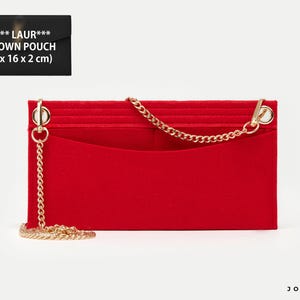 May include: A bright red rectangular felt pouch featuring a gold-tone metal chain strap attached to two circular rings. The pouch has an open top pocket and is labeled SAI** LAUR*** UPTOWN POUCH with dimensions of 27 x 16 x 2 cm printed on the item.