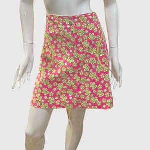 Puede incluir: Una minifalda rosa con estampado floral verde y ribete amarillo. La falda tiene cierre de botón y bolsillos.