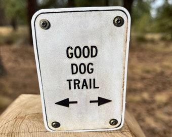 May include: A white sign with black text and arrows that reads "GOOD DOG TRAIL". The sign is attached to a wooden post.