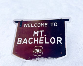 May include: A brown and white sign with the text "Welcome to Mt. Bachelor" and a U.S. Forest Service logo.