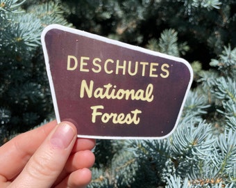 May include: A sticker with a dark background and white border. The text "Deschutes National Forest" is written in yellow.