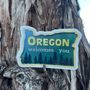 May include: A green and yellow sticker shaped like the state of Oregon with the text "Oregon welcomes you" and a row of evergreen trees along the bottom.