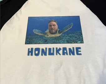 May include: White and black raglan-style t-shirt featuring a humorous image of a person's face superimposed on a sea turtle, set against an underwater backdrop. The word "HONUKANE" is printed in blue below the image.
